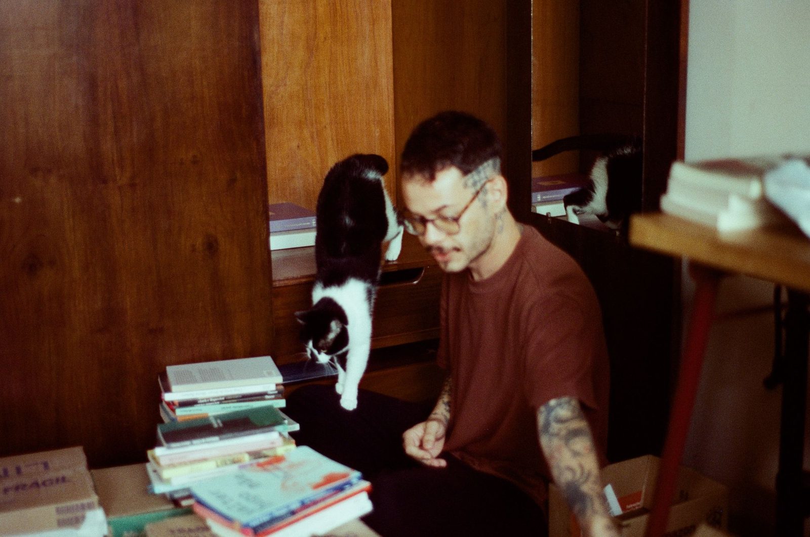 Valmir Jr no ateliê, entre pilhas de livros, com um gato preto e branco descendo da estante